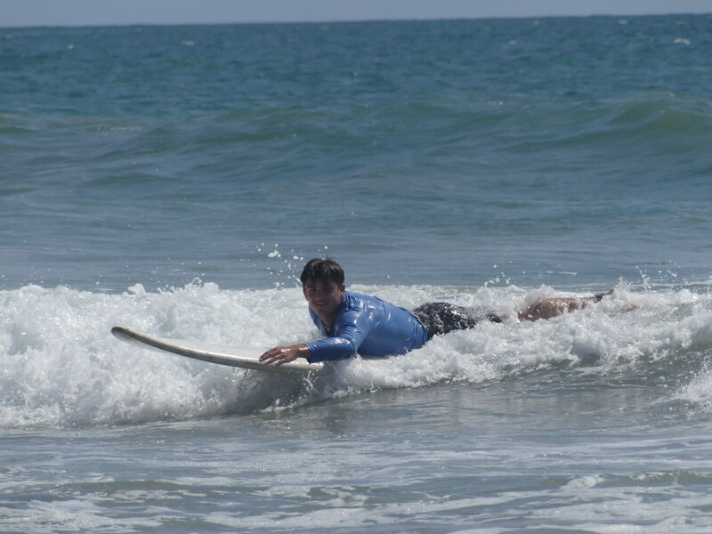 A person is lying on a surfboard in the ocean, paddling with their hands. They are wearing a blue long-sleeved shirt and dark shorts. The surfboard is light-colored, and the water is choppy with whitecaps. The sky is visible in the background and appears clear. The person seems to be enjoying the activity of surfing or attempting to catch a wave.